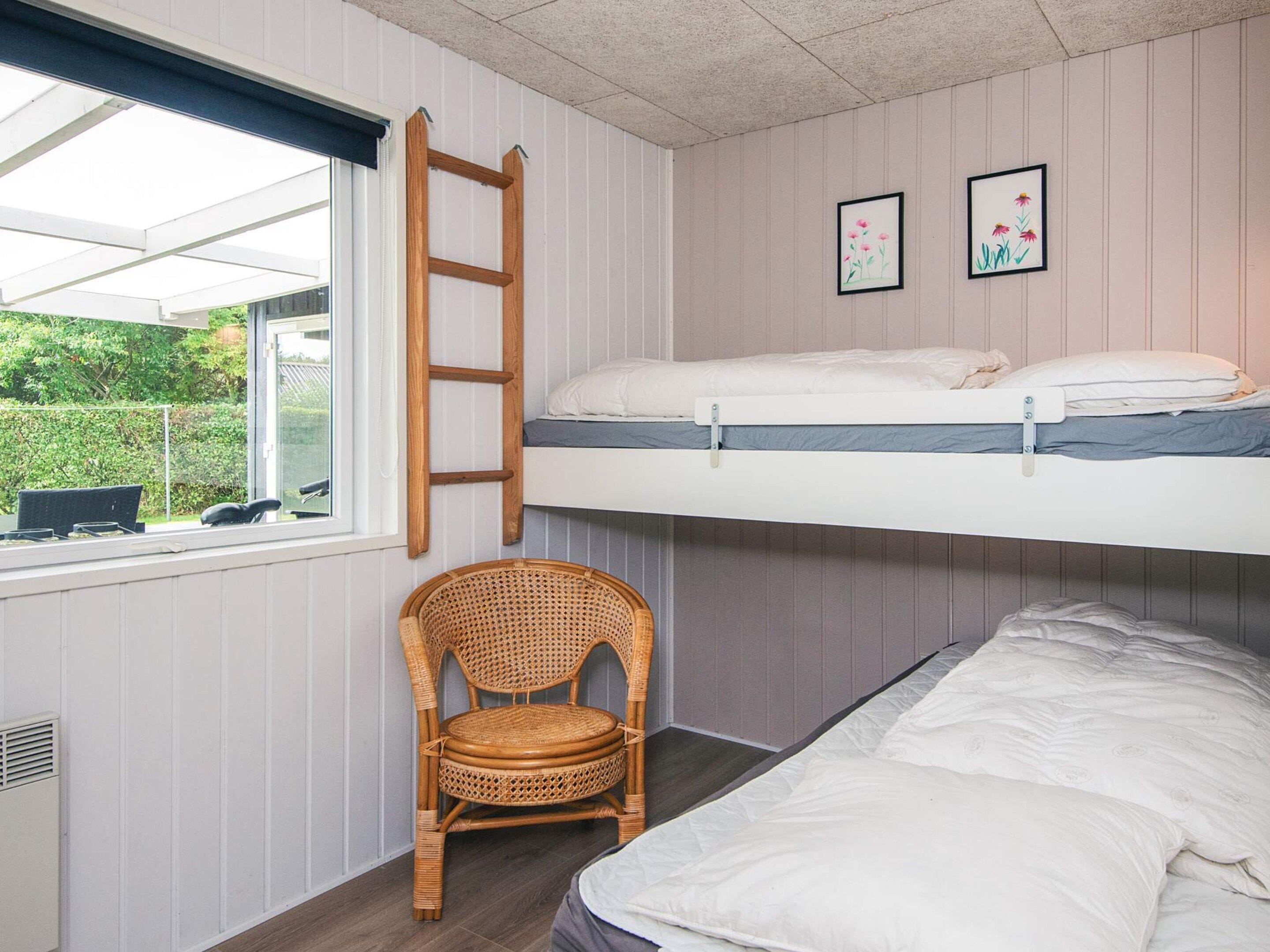 Una habitación con una cama litera, una silla de ratán, una ventana con persianas y dos cuadros en la pared.