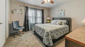 A bedroom with a bed, a desk, a chair, a ceiling fan, a window with curtains, and a wall shelf.