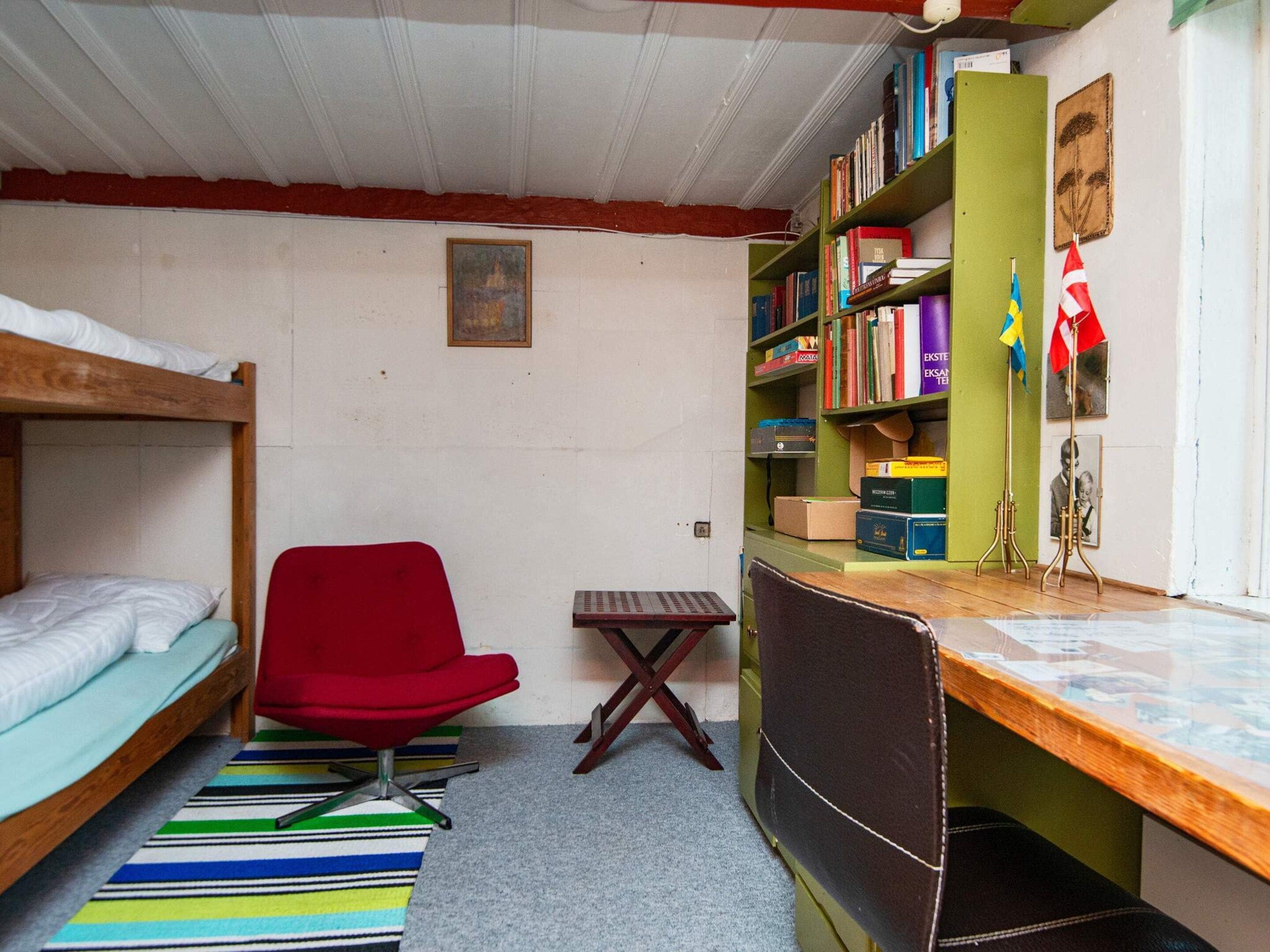 Une chambre de dortoir avec des lits superposés, une chaise rouge, un bureau avec un ordinateur et une bibliothèque.