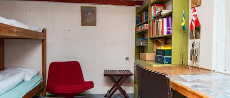 A dormitory room with bunk beds, a red chair, a desk with a computer, and a bookshelf.
