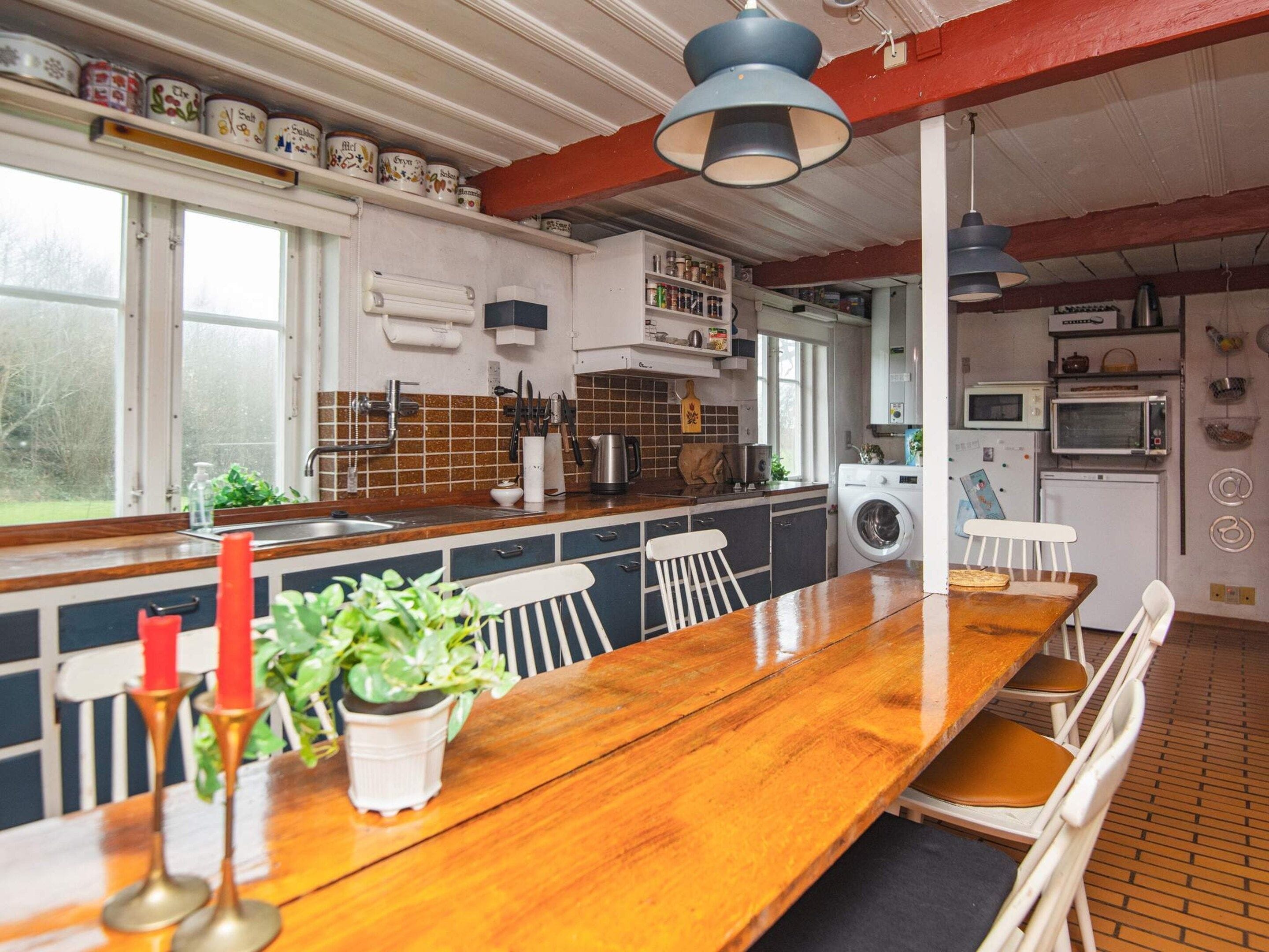 Una cocina con una mesa de madera, sillas, un lavabo, una lavadora y diversos utensilios de cocina.