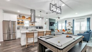 Une cuisine moderne dotée d’un îlot central intégré, d’appareils électroménagers en acier inoxydable et d’une table de billard dans un espace de vie agrémenté d’un canapé bleu et d’une grande fenêtre.