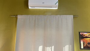 Una stanza con un condizionatore bianco montato su un muro giallo, una tenda bianca e un quadro con barche sull'acqua.