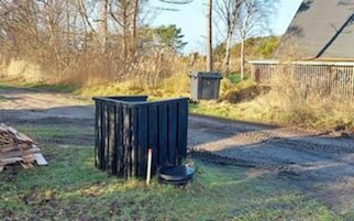 Entorno rural con un bote de basura negro y un recipiente más pequeño, rodeado de hierba y árboles.