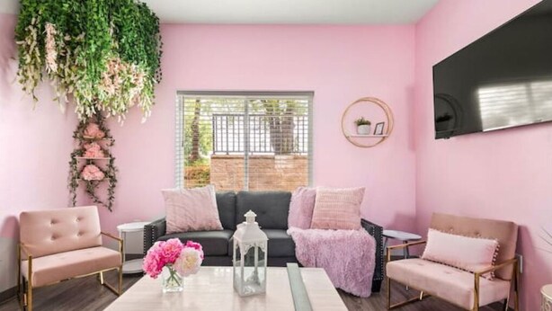 Ett mysigt vardagsrum med en rosa vägg, en svart soffa och två rosa fåtöljer. Ett vitt bord med en vas med blommor står i mitten.