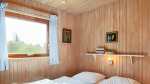 Ein Zimmer mit Holzvertäfelung, einem Bett, einem Fenster mit Vorhängen und zwei an der Wand befestigten Regalen mit Büchern.