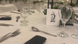Uma mesa de jantar posta com cartões de lugar numerados, copos de vinho, talheres e um guardanapo com um logotipo.