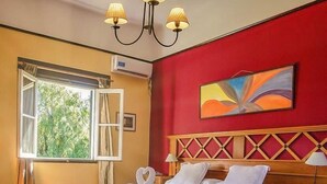 Quarto de hotel com uma cama, mesa de cabeceira, luminária, quadro, aparelho de ar condicionado e janela com cortinas.