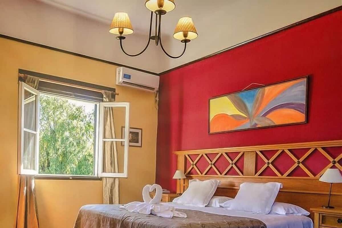 Une chambre d’hôtel avec un lit, une table de chevet, une lampe, un tableau, un appareil de climatisation et une fenêtre avec des rideaux.