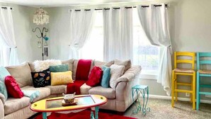 Una sala de estar con un sofá beige, cojines coloridos, un tapete rojo y una mesa de centro amarilla.