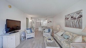 A spacious living area with a flat-screen TV, white furniture, and a dining area in the background.