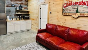 A cozy room with a red leather couch, wooden walls, a kitchen area with white cabinets, and a sign for "The Grand Pajoni" country theater. - Knotty Squirrel Unit 9 Standard King Suite (Mountain View)