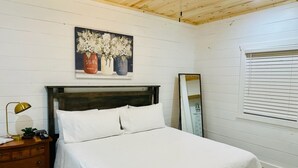 A bedroom with a wooden ceiling, a bed with white bedding, a nightstand with a lamp, a dresser, a painting of flowers, and a window with blinds. - Knotty Squirrel Unit 9 Standard King Suite (Mountain View)