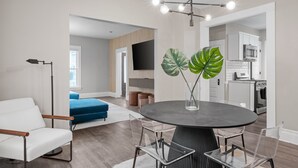 A modern living room with a round dining table, clear chairs, a white sofa, and a large green plant.
