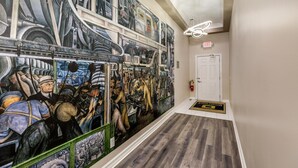 Un couloir orné d’une grande fresque représentant des scènes industrielles, une porte blanche avec un panneau indiquant la sortie, et un luminaire moderne.