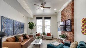 Un salon moderne avec un canapé en cuir marron, une table basse blanche et des fauteuils turquoise. Il y a deux fenêtres avec des stores noirs, un mur en briques et un ventilateur de plafond.
