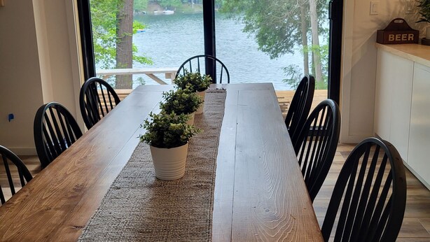 A dining area with a wooden table, black chairs, a woven placemat, and a potted plant. A large window offers a view of a lake and trees. - Cozy Lakeside Living (Verona)