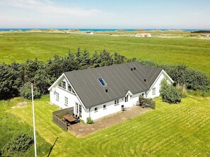 A white two-story house with a dark roof, surrounded by a well-maintained lawn and trees, with a view of the ocean in the background. - 10 Person Holiday Home in Thisted (Thisted)