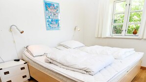 A neatly made bed with white bedding, a bedside table with a lamp, and a window with a view of greenery.