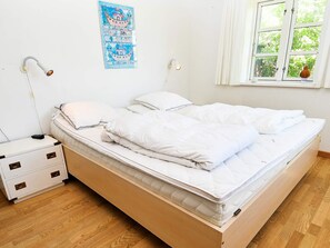 A neatly made bed with white bedding, a bedside table with a lamp, and a window with a view of greenery.