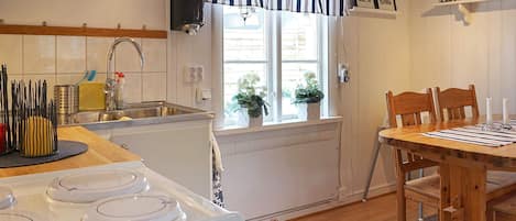 Une cuisine équipée d’une cuisinière, d’un évier et d’une fenêtre avec des rideaux. On y trouve une table à manger avec des chaises et une petite étagère sur laquelle sont posés divers objets.