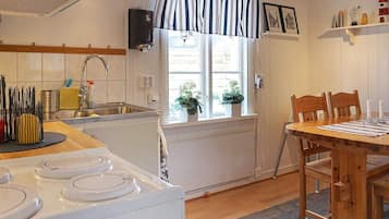 A kitchen with a stove, sink, and window with curtains. There is a dining table with chairs and a small shelf with items.