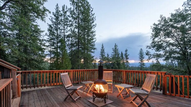 A wooden deck with a fire pit and two chairs, surrounded by a wooden railing and tall trees.