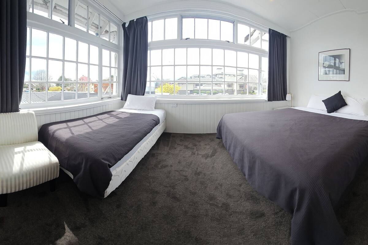 Une chambre d’hôtel avec deux lits, de grandes fenêtres, un radiateur, une table de chevet et un tableau encadré au mur.