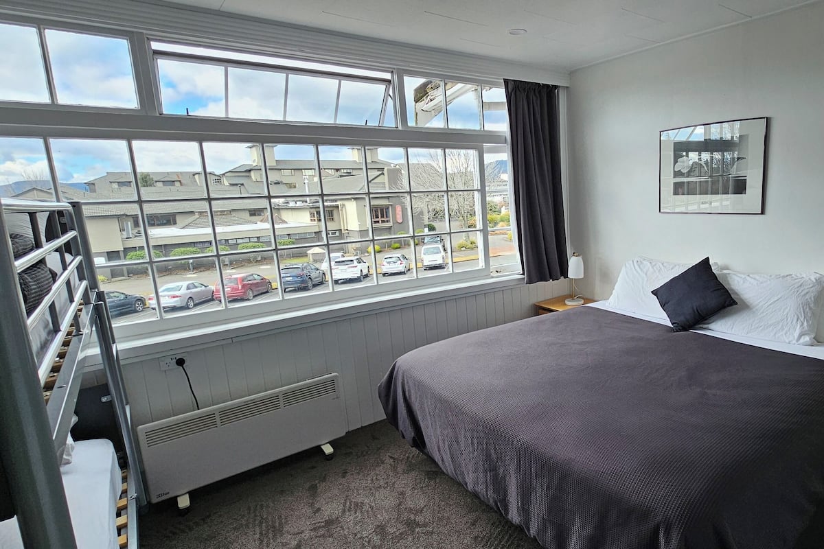 Une chambre d’hôtel avec une grande fenêtre, un lit recouvert d’une courtepointe sombre, une table de chevet avec une lampe et une petite table de nuit.