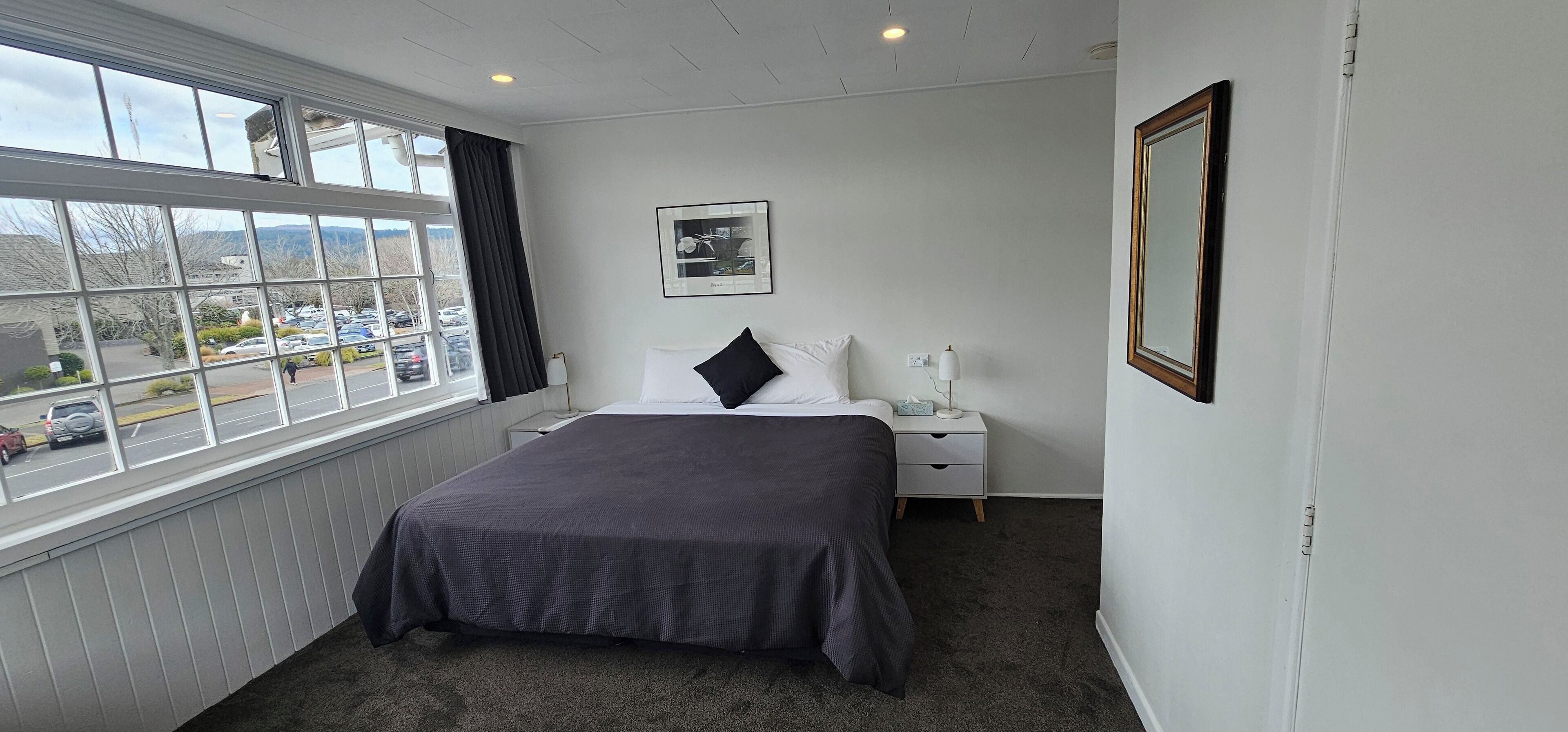 A hotel room with a large window, a bed with a dark bedspread, a nightstand with a lamp, and a framed picture on the wall.