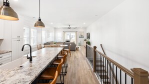 A modern kitchen with a marble countertop, bar stools, pendant lighting, and a staircase with a wooden railing. - Pearl Paradise: with elevator & golf cart included! 2 blocks to beach.  (Carolina Beach)