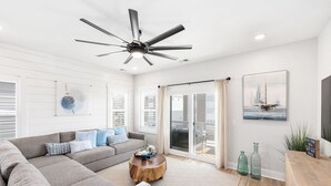 A modern living room with a sectional sofa, a round wooden coffee table, a ceiling fan, and a large window with curtains. - Pearl Paradise: with elevator & golf cart included! 2 blocks to beach.  (Carolina Beach)