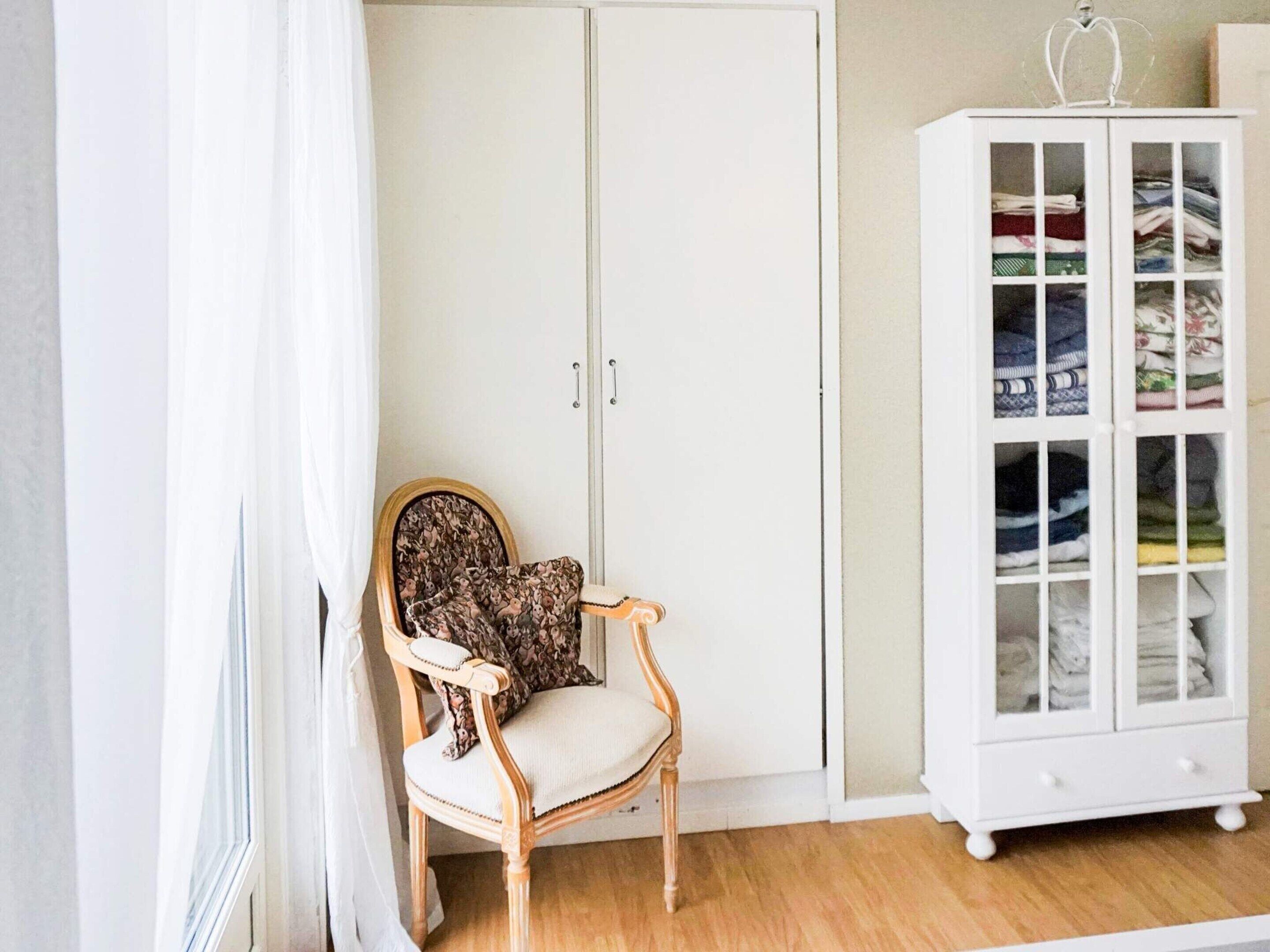 A room with a wooden chair, a wardrobe, and a cabinet with folded clothes.