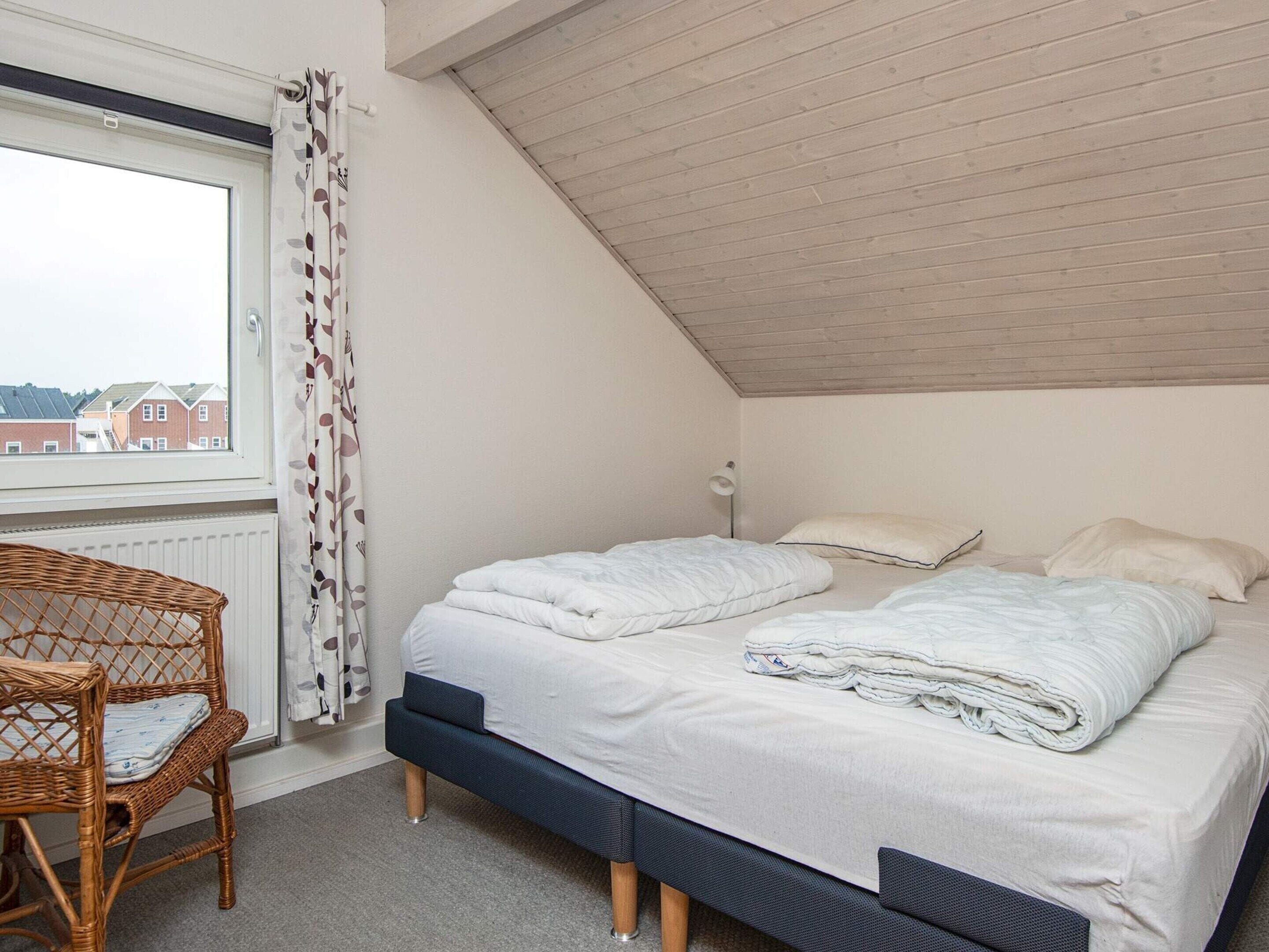 Ein Schlafzimmer mit schrägem Dach, einem großen Fenster mit Vorhängen, einem Bett mit weißer Bettwäsche, einer Nachttischlampe und einem Korbstuhl.