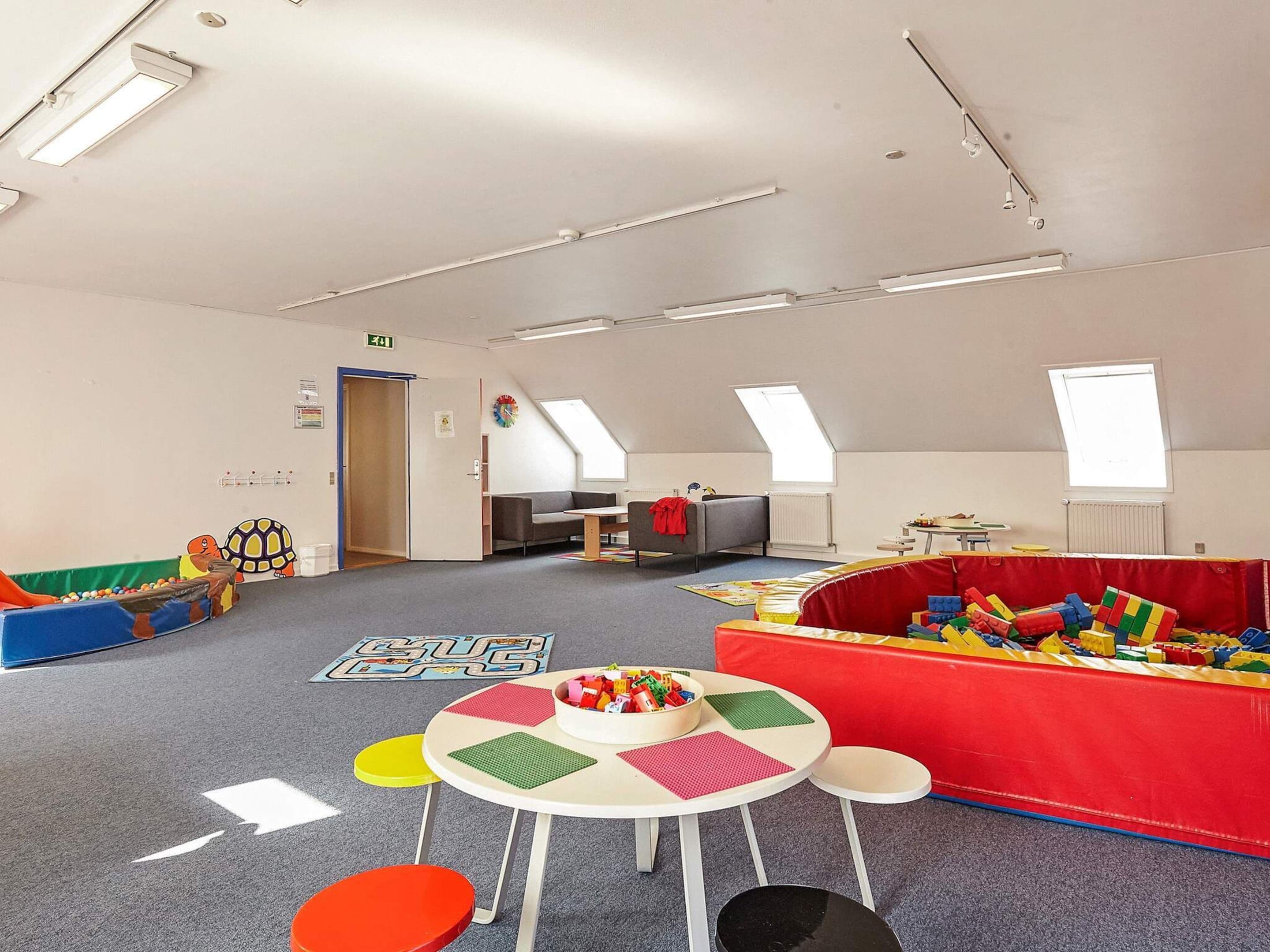 A spacious playroom with a high ceiling, skylights, and a variety of play structures including a soft play area and a table with colorful mats.