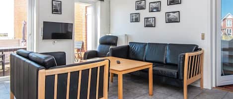 A modern living room with a sofa, armchair, and coffee table. There is a TV mounted on the wall and several framed pictures.
