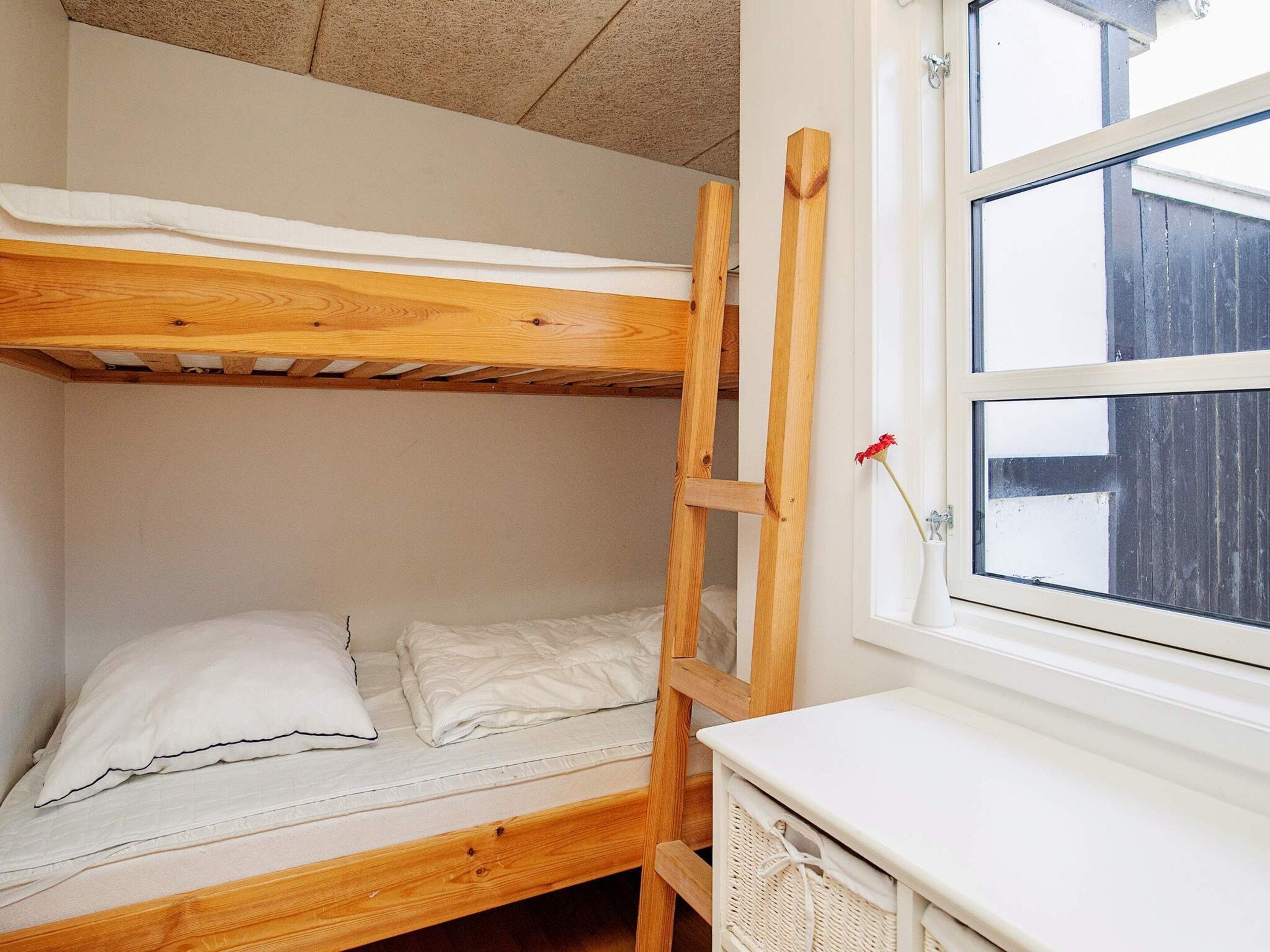 Ein Stockbett mit einer Holzleiter, ein weißer Nachttisch und ein Fenster mit Blick auf einen Holenzaun.
