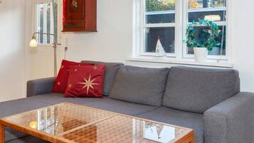 A modern living room with a grey sofa, red and gold cushions, a glass coffee table, a wooden cabinet, and a window with a view of the outside.