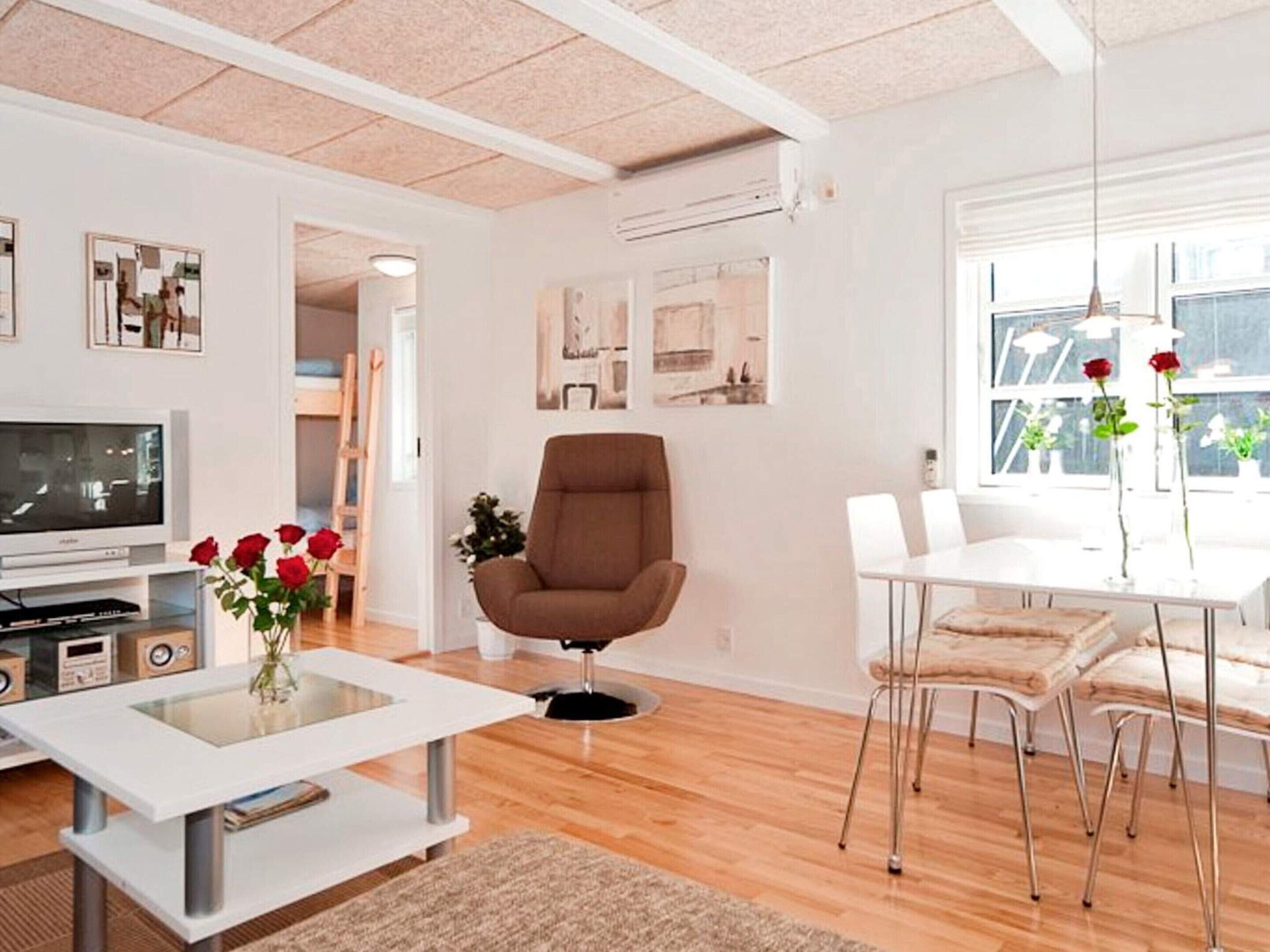 Una sala de estar moderna con televisor de pantalla plana, un sillón marrón, una mesa de centro de vidrio y un comedor con sillas blancas.