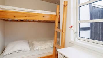 Una cama litera con escalera de madera, colchón blanco y almohada blanca. Se ve una ventana con vista a una cerca de madera.