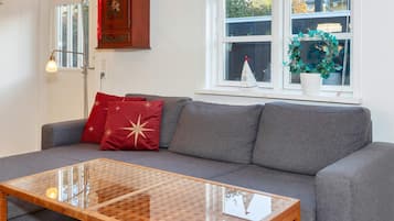 A living room with a grey sofa, red and gold cushions, a glass coffee table, a wooden cabinet, and a window with a view of trees and a building.