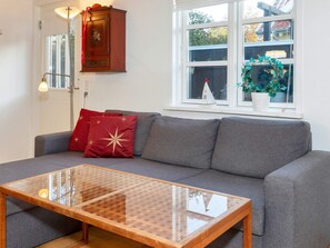 A living room with a grey sofa, red and gold cushions, a glass coffee table, a wooden cabinet, and a window with a view of trees and a building. - 4 Star Holiday Home in Skagen (Skagen)