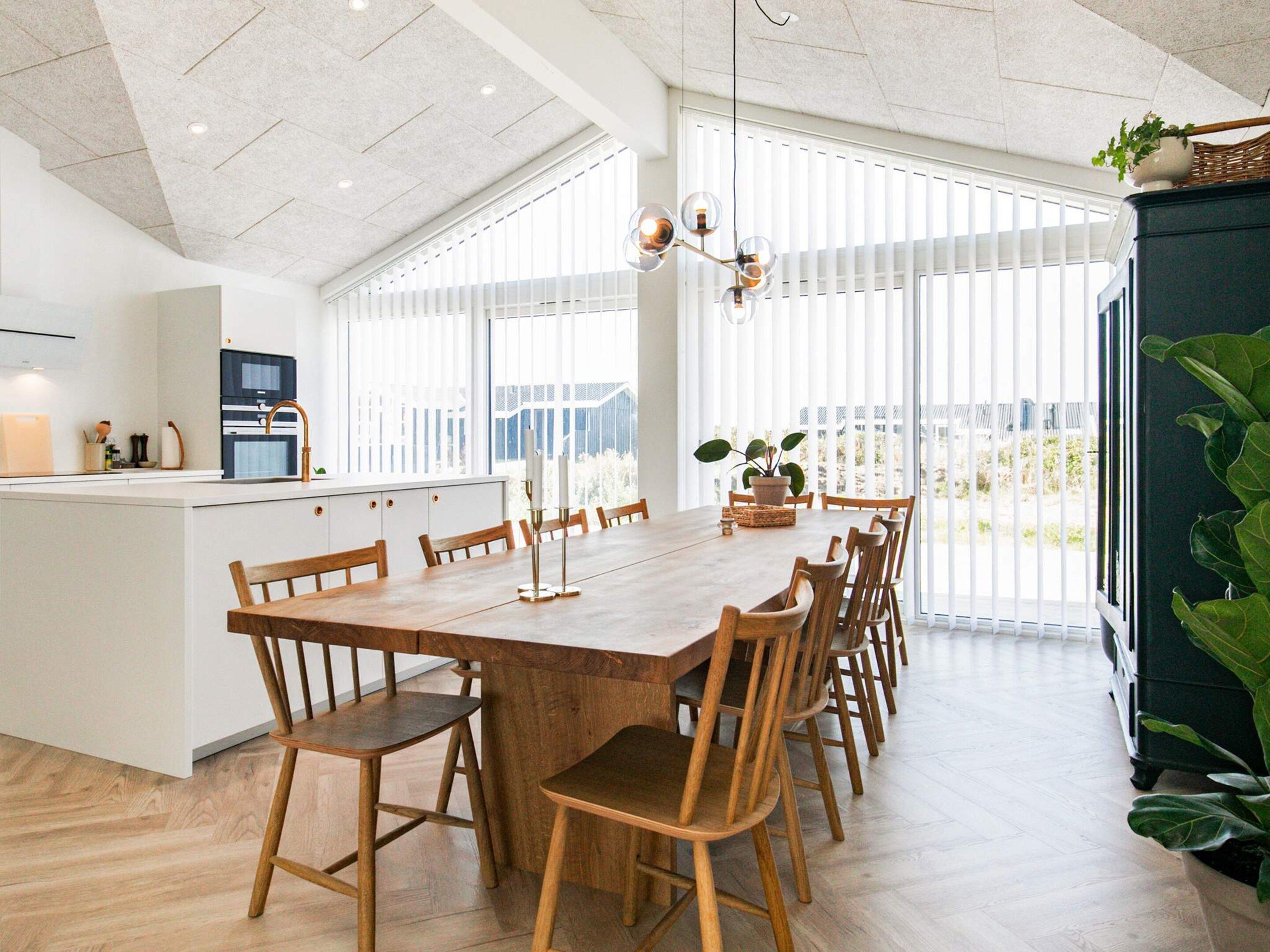Un comedor amplio con una larga mesa de madera y sillas, una cocina al fondo y ventanas grandes que dejan entrar luz natural.