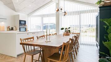 Un comedor amplio con una larga mesa de madera y sillas, una cocina al fondo y ventanas grandes que dejan entrar luz natural.