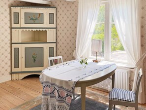 A room with a wooden floor, a round table with a tablecloth, a chair, a cabinet with floral designs, and a window with curtains.