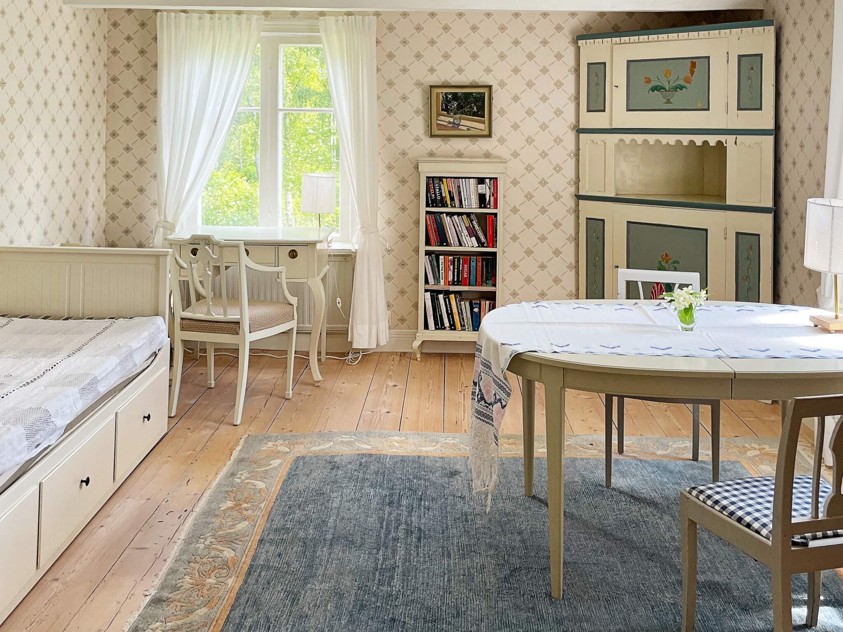 A room with a bed, a desk, a chair, a bookshelf, and a table with a vase of flowers.