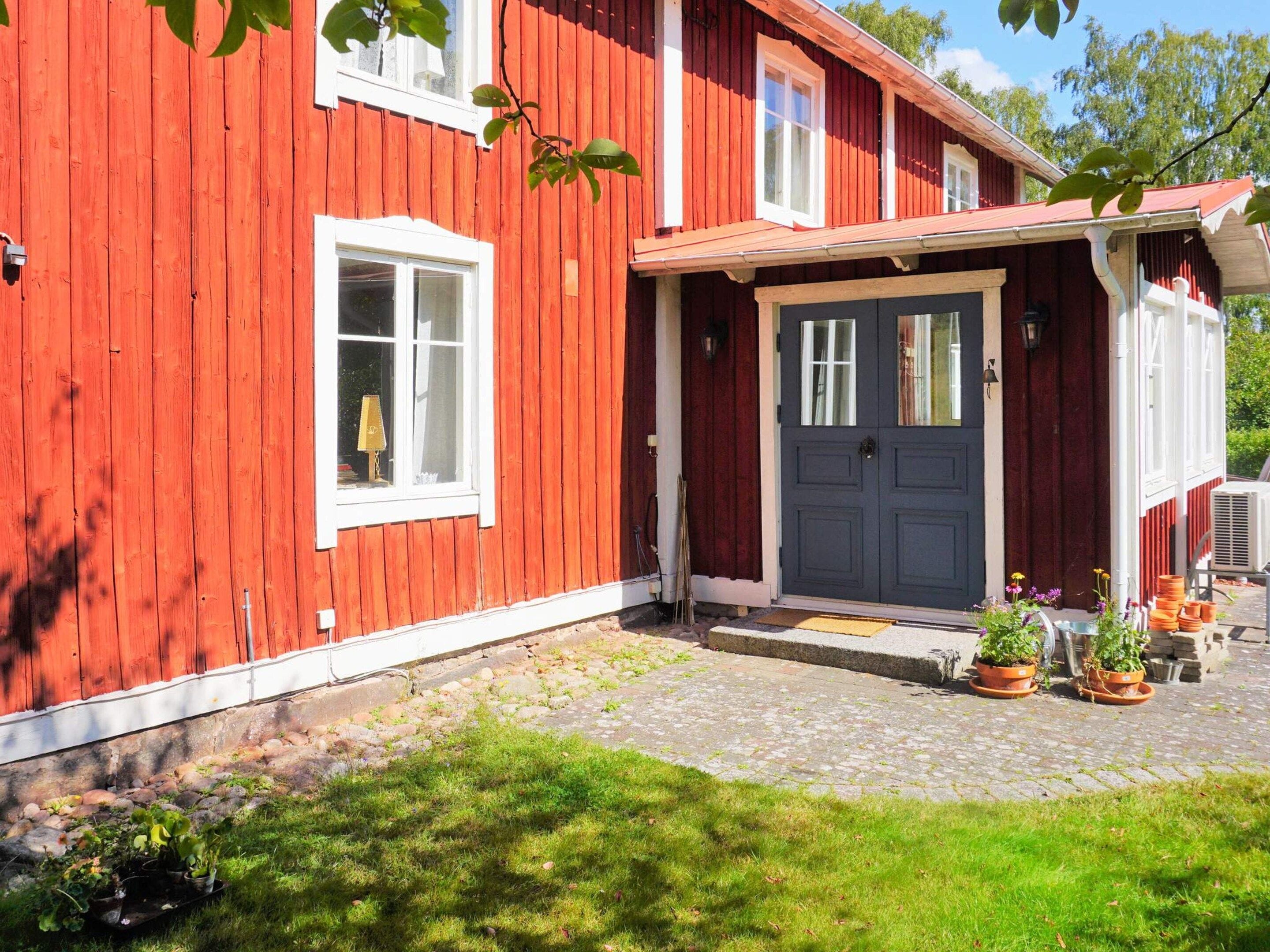 A red wooden house with a dark blue door, white windows, and a small porch area with potted plants.