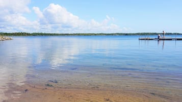 Un lago tranquilo con cielo despejado, orilla arenosa y un muelle con gente.