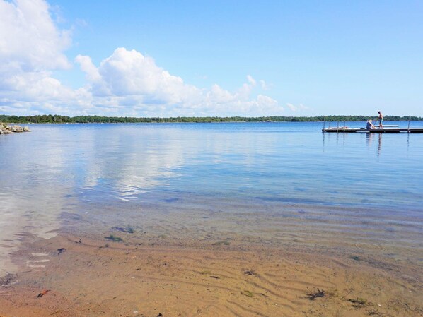 A calm lake with a clear sky, a sandy shore, and a dock with people.