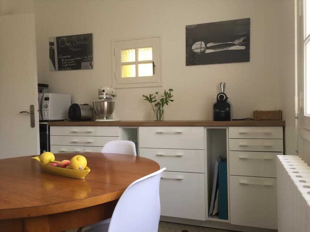 A compact kitchen with a wooden table, white chairs, a fruit bowl, a kettle, a coffee maker, and a small window.
